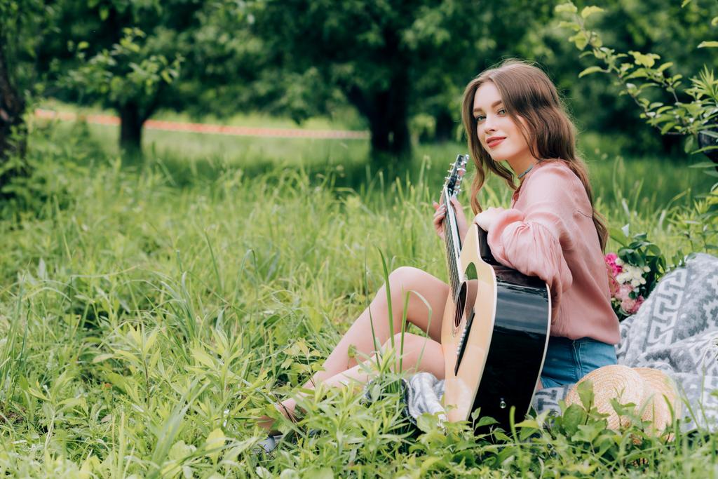 Widok z boku młodej kobiety z gitarą akustyczną na koc z bukietem kwiatów w parku - Zdjęcie, obraz
