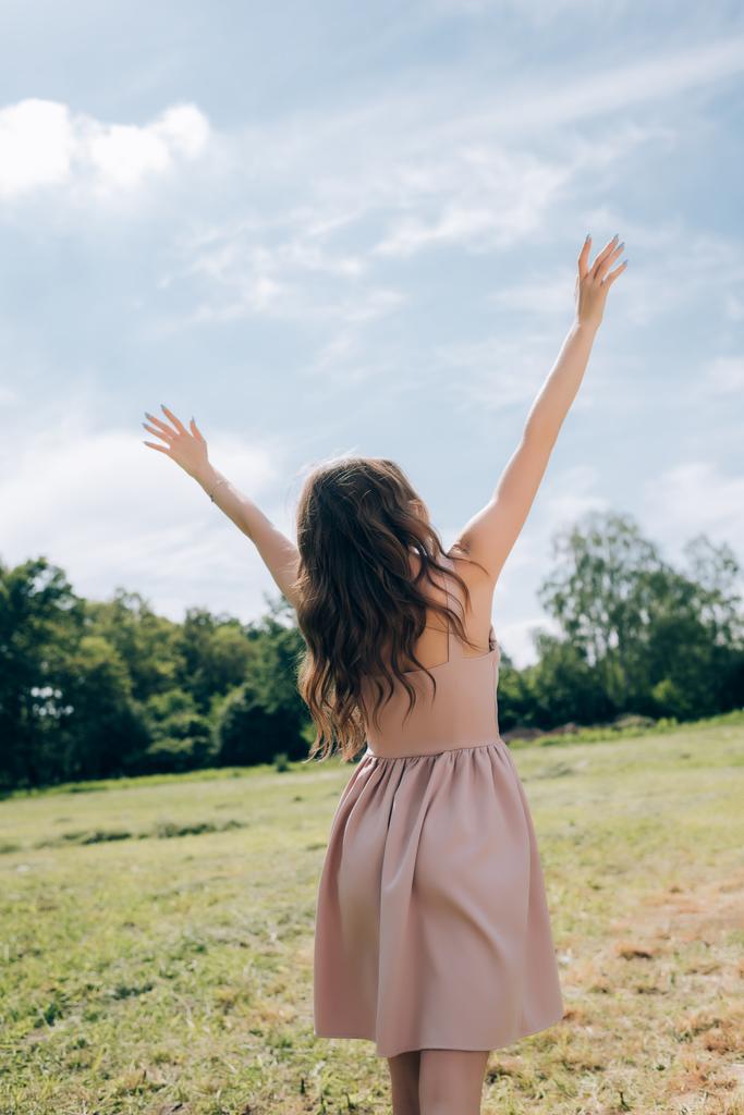 rear view of woman in stylish dress with outstretched arms standing in meadow with blue sky on background - Photo, Image