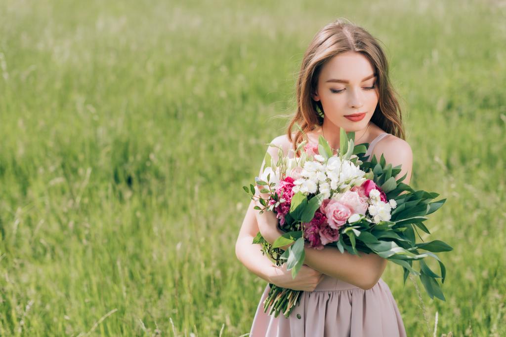 Porträt einer schönen nachdenklichen Frau, die einen Blumenstrauß in der Hand hält, während sie allein auf dem Feld steht - Foto, Bild