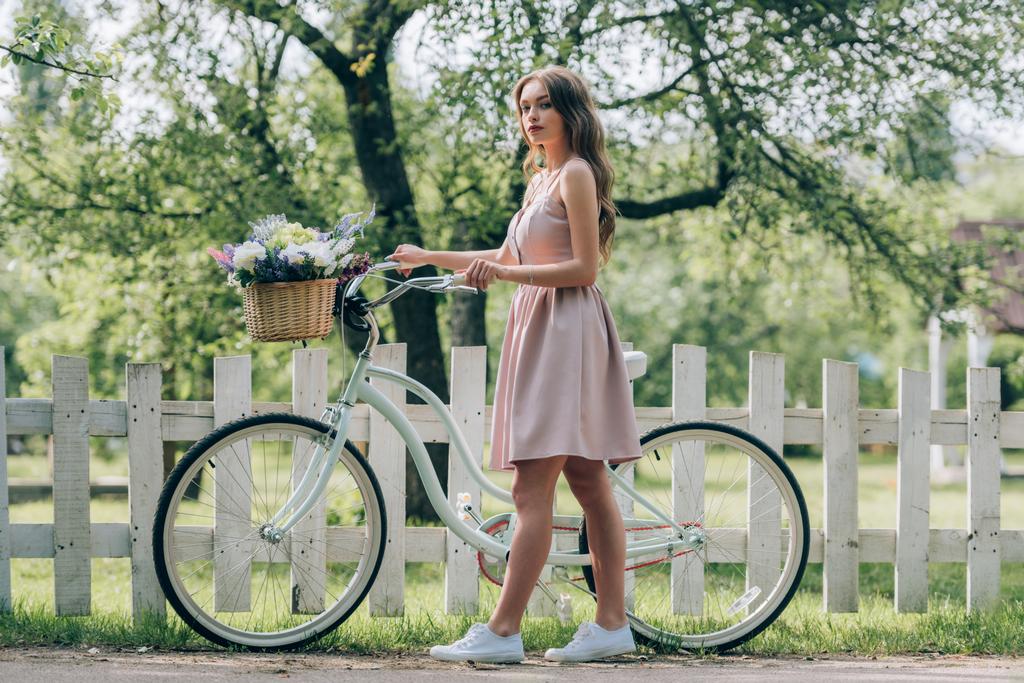 vue latérale de la jeune femme en robe avec vélo rétro avec panier en osier plein de fleurs à la campagne
 - Photo, image