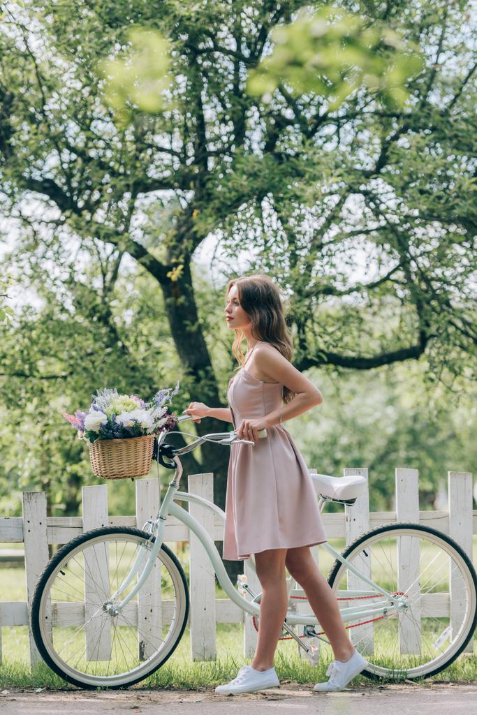 vue latérale de la jeune femme en robe avec vélo rétro avec panier en osier plein de fleurs à la campagne
 - Photo, image