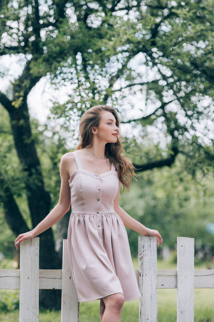 nachdenklich schöne Frau in stilvollem Kleid steht am weißen Zaun auf dem Land - Foto, Bild