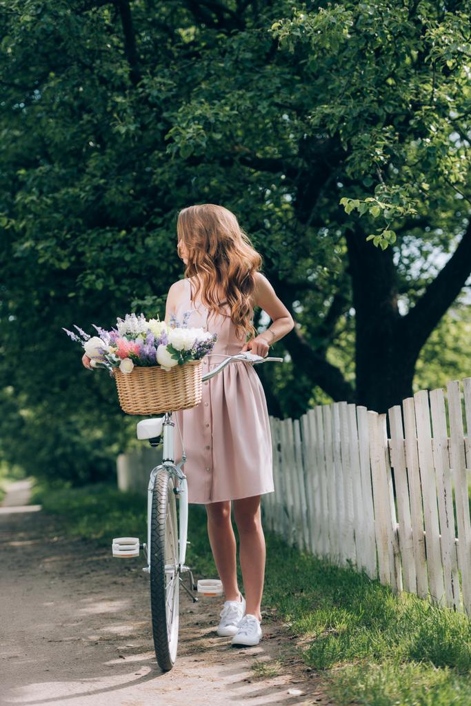 young woman in dress with retro bicycle with wicker basket full of flowers at countryside - Photo, Image