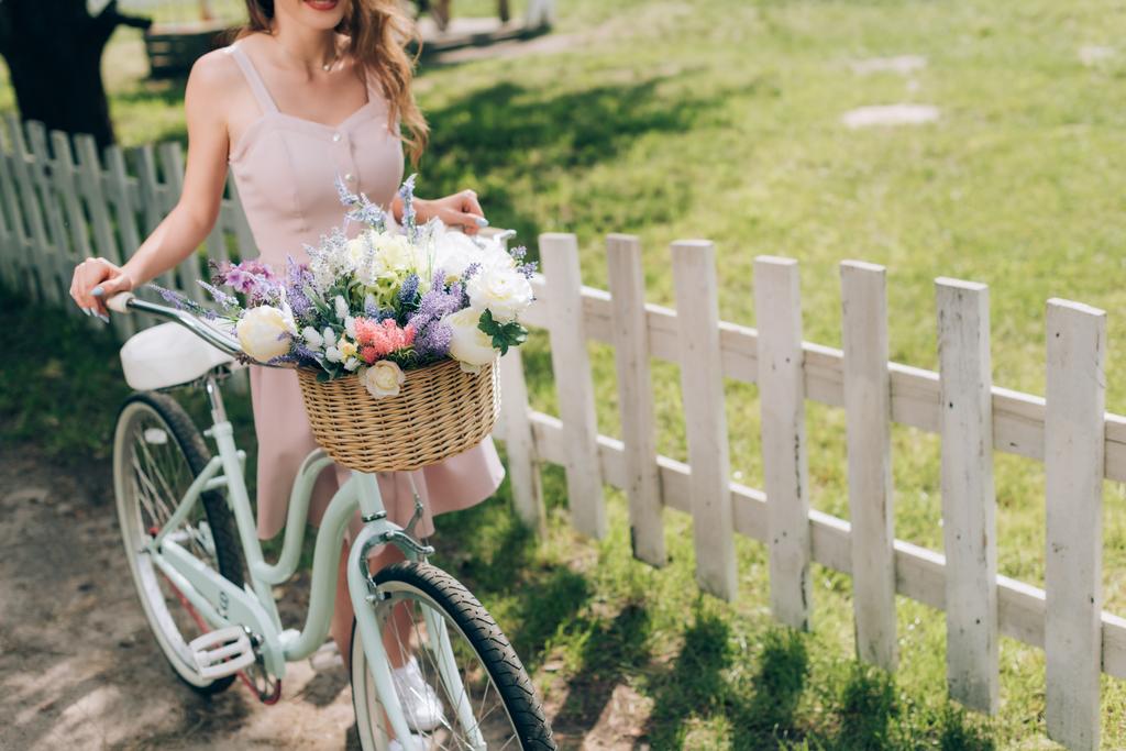 partial view of woman in stylish dress with retro bicycle with wicker basket full of flowers at countryside - Photo, Image