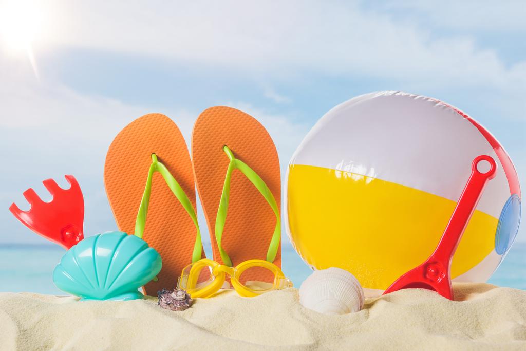 Slippers met strandbal en speelgoed in zand op blauwe hemelachtergrond - Foto, afbeelding