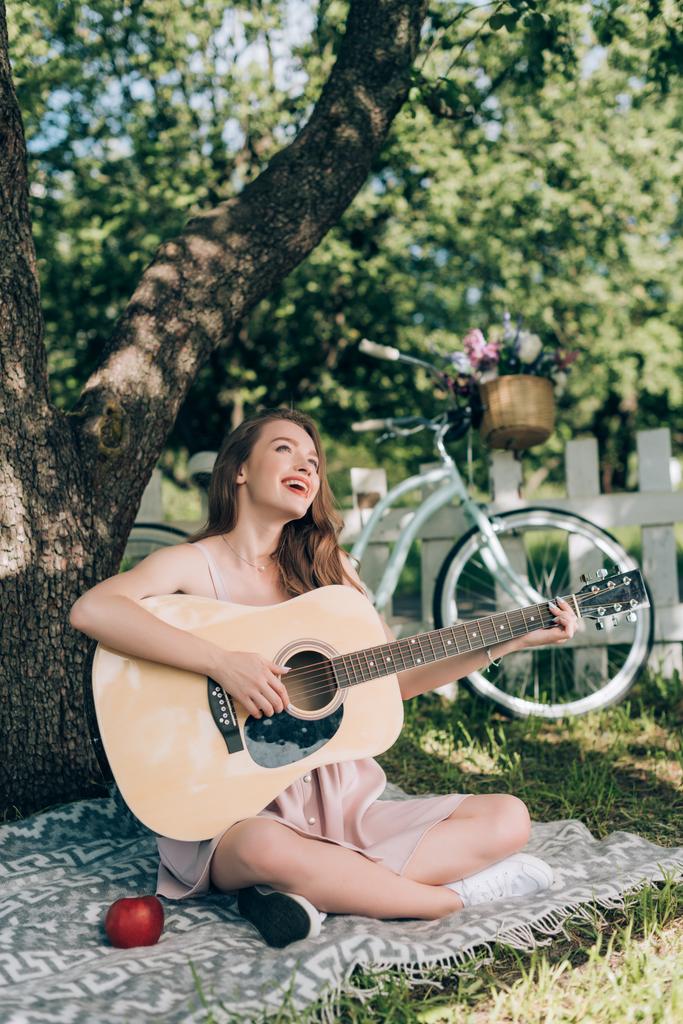 atrakcyjna młoda kobieta gra na gitarze akustycznej podczas odpoczynku na koc pod drzewem na wsi - Zdjęcie, obraz