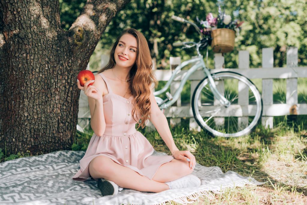 Улыбающаяся задумчивая женщина с спелым яблоком в руке отдыхает на одеяле под деревом в сельской местности
 - Фото, изображение