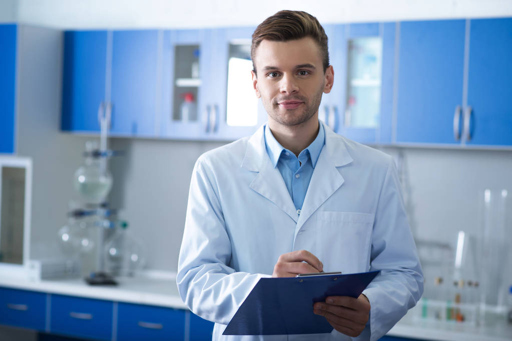 Best employers. Adult handsome self-confident scientist standing in the lab looking straight and keeping his notebook. - Photo, Image