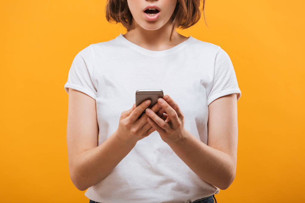 Обрізана фотографія збудженої молодої жінки, що стоїть ізольовано на жовтому фоні спілкування на мобільному телефоні
. - Фото, зображення
