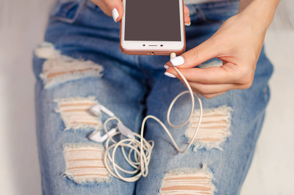 Mulher de jeans olha para celular smartphone e fones de ouvido
 - Foto, Imagem