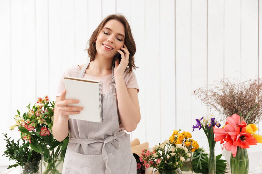 Jovem florista mulher de pé perto da mesa com flores diferentes em estúdio e chamando clientes com notas na mão
 - Foto, Imagem
