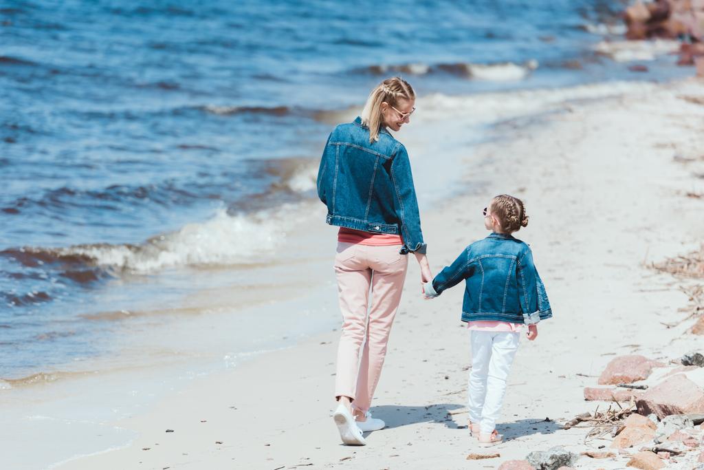 rear view of mom and daughter holding hands and walking on sea shore - Photo, Image