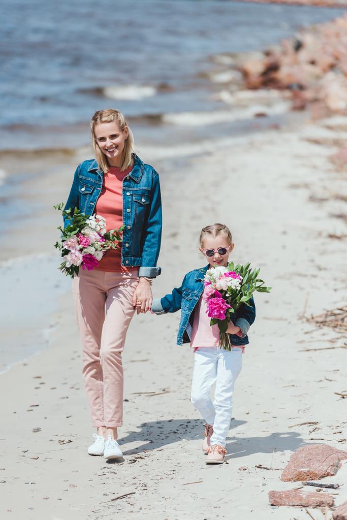 привлекательная мать и счастливая дочь с пионерскими букетами, держащимися за руки и гуляющими по берегу моря
 - Фото, изображение