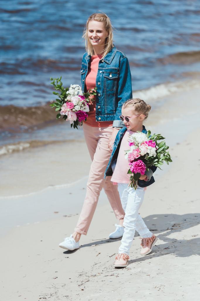 mother and daughter walking with peony bouquets on sea shore - Photo, Image