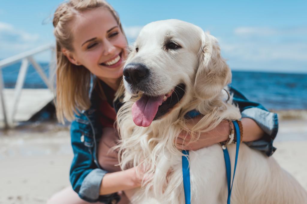улыбающаяся блондинка, сидящая с дружелюбной собакой на берегу моря
 - Фото, изображение