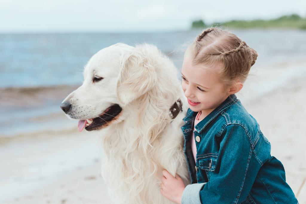  αξιολάτρευτο παιδί αγκαλιάζει golden retriever σκύλου στην ακτή θάλασσας - Φωτογραφία, εικόνα