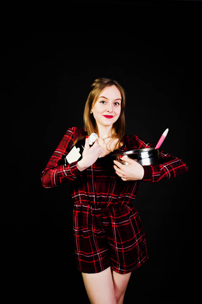 Mladá hospodyňka v kostkované šaty s pánev a kuchyňské lžíce a láhev izolované na černém pozadí. - Fotografie, Obrázek