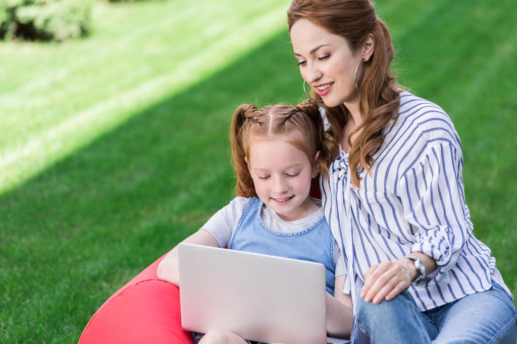 portrait of mother and daughter using laptop while resting on bag chair together - Photo, Image