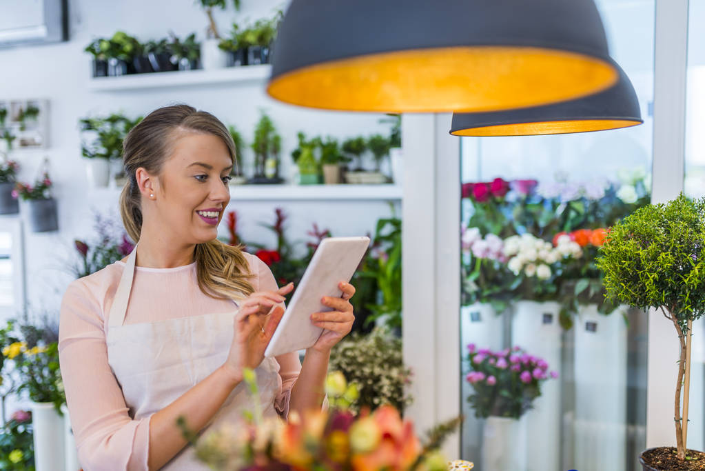 Young owner of flower shop taking orders online by tablet. Small business. Beautiful female florist with tablet in flower shop. Florist using interface of online flower shop - Photo, Image