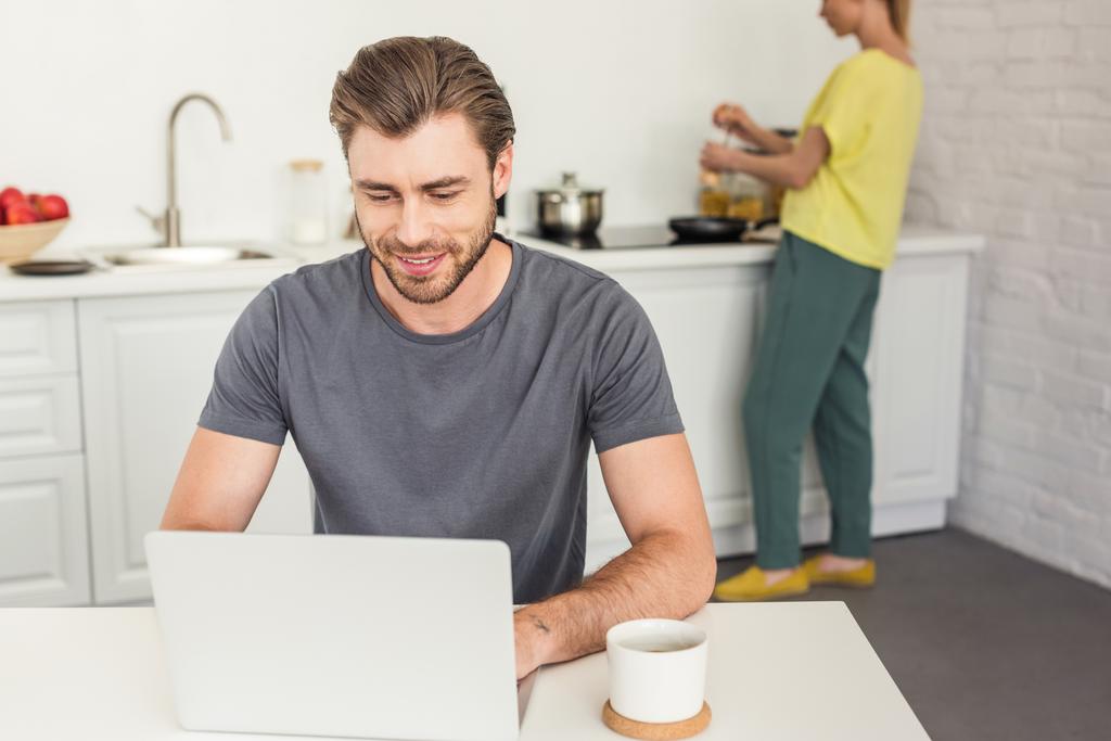 sorrindo jovem trabalhando no laptop e sua namorada cozinhar atrás na cozinha
  - Foto, Imagem