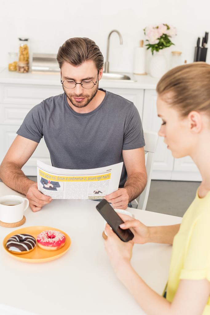 Zijaanzicht van vrouw met behulp van smartphone en vriendje lezen van de krant aan tafel met koffie kopjes en donuts op keuken  - Foto, afbeelding