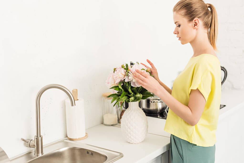 boční pohled na mladou ženu dojemné kytice pivoněk v kuchyni - Fotografie, Obrázek