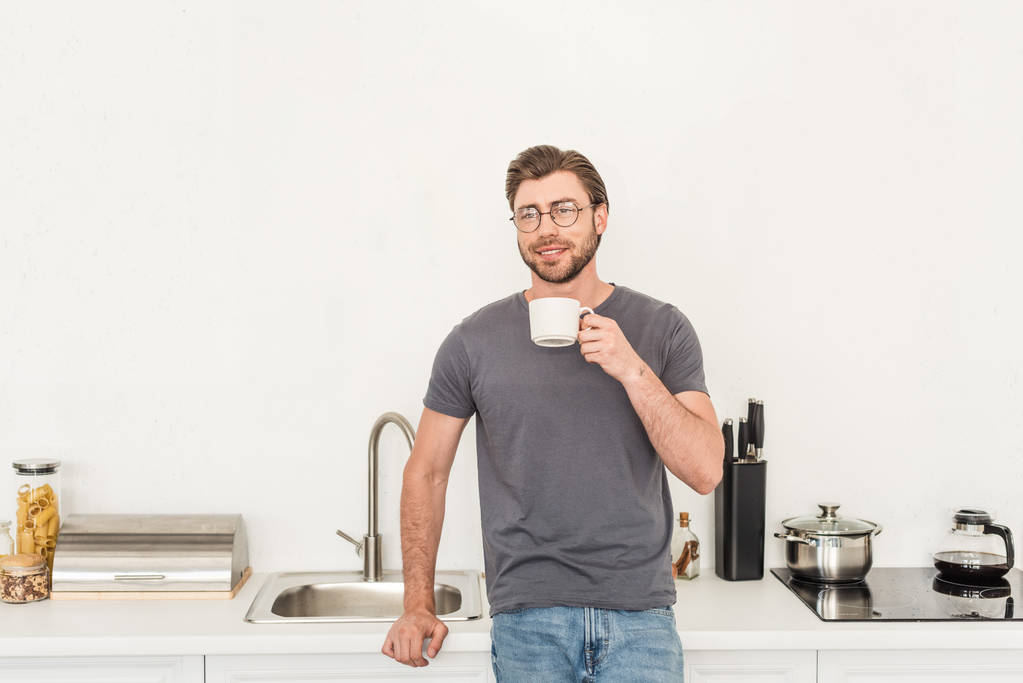 キッチンでコーヒーを飲んで眼鏡の若い男を笑顔のフロント ビュー  - 写真・画像