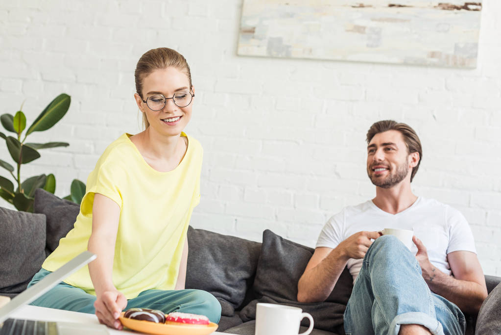 mladý usmívající se pár s snídaně s kávou a koblihy na gauči doma - Fotografie, Obrázek