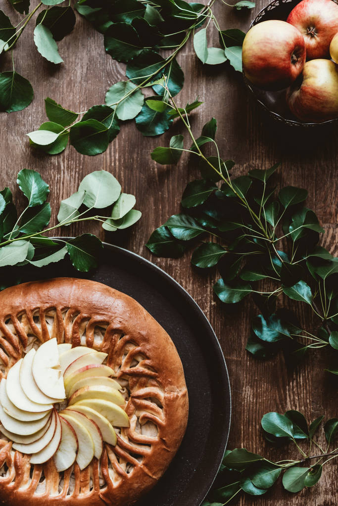 Blick von oben auf hausgemachten Apfelkuchen und frische Äpfel mit grünen Blättern auf Holztischplatte - Foto, Bild