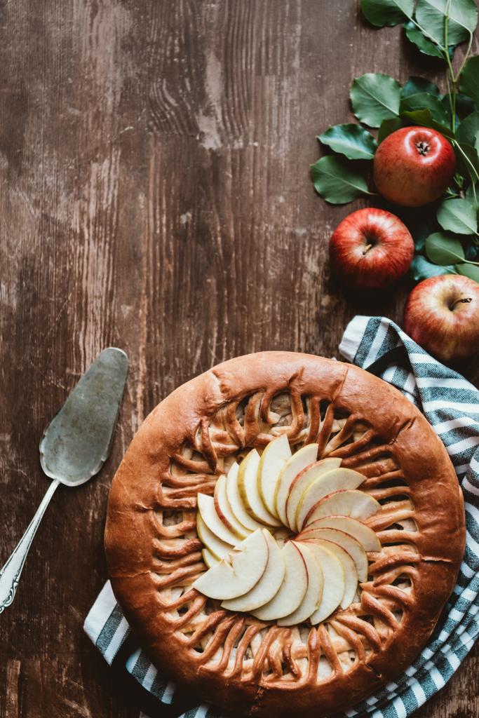 Draufsicht auf arrangierten Apfelkuchen, Kuchenserver und frische Äpfel mit grünen Blättern auf Holzoberfläche - Foto, Bild