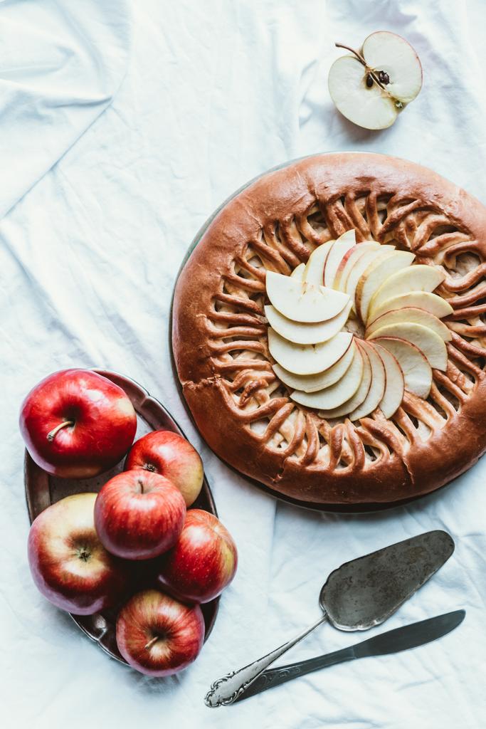 Draufsicht auf frische Äpfel, Messer und hausgemachten Apfelkuchen auf weißer Tischdecke - Foto, Bild