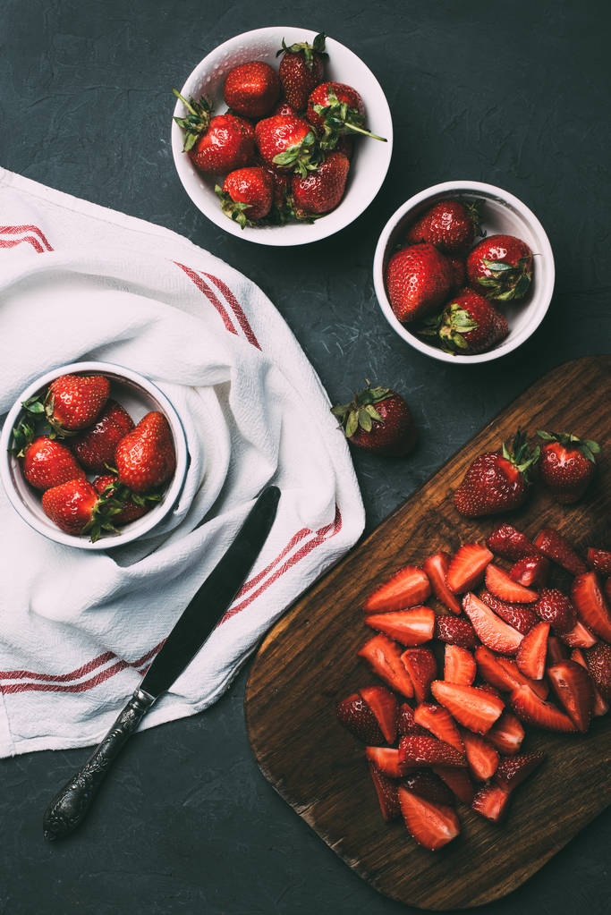 top view or ripe whole strawberries in bowls and sliced strawberries on wooden cutting board on black  - Photo, Image