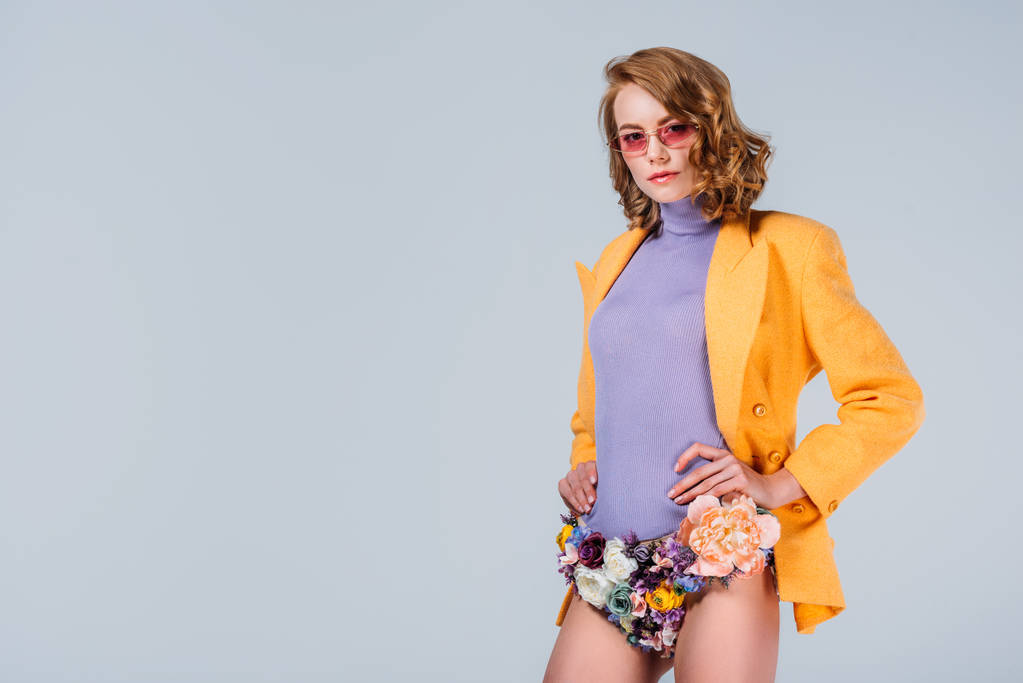 beautiful young woman in sunglasses and panties made of flowers standing with hands on waist and looking at camera isolated on grey - Photo, Image