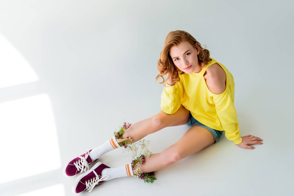 high angle view of beautiful girl looking at camera while sitting with flowers in socks on grey - Photo, Image
