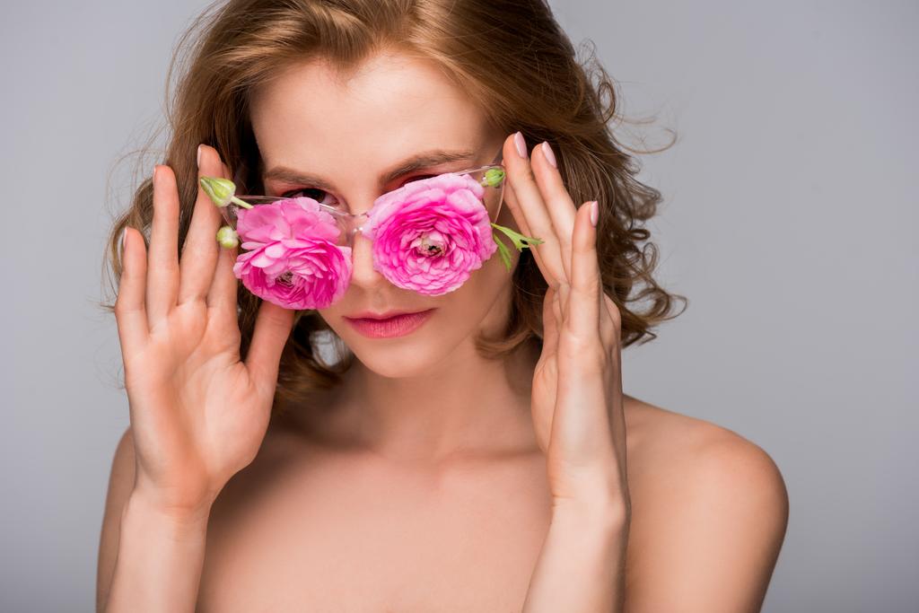 schöne junge nackte Frau, die ihre Brille mit Blumen anpasst und isoliert auf grau in die Kamera schaut - Foto, Bild