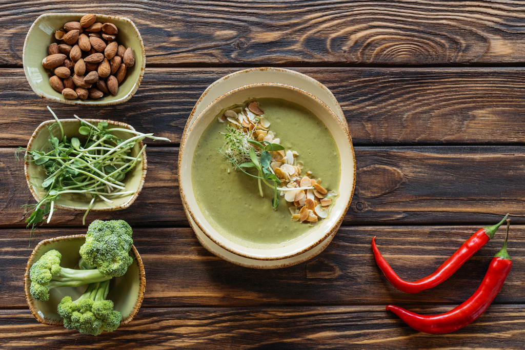 bovenaanzicht van gearrangeerde vegetarische crème soep, spruiten, amandelen en verse broccoli in kommen op houten oppervlak - Foto, afbeelding