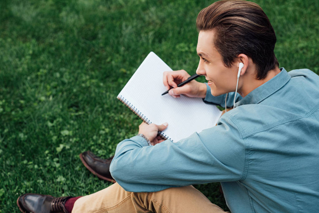 high angle view of smiling young man in earphones taking notes in blank notebook and looking away while sitting on grass - Photo, Image