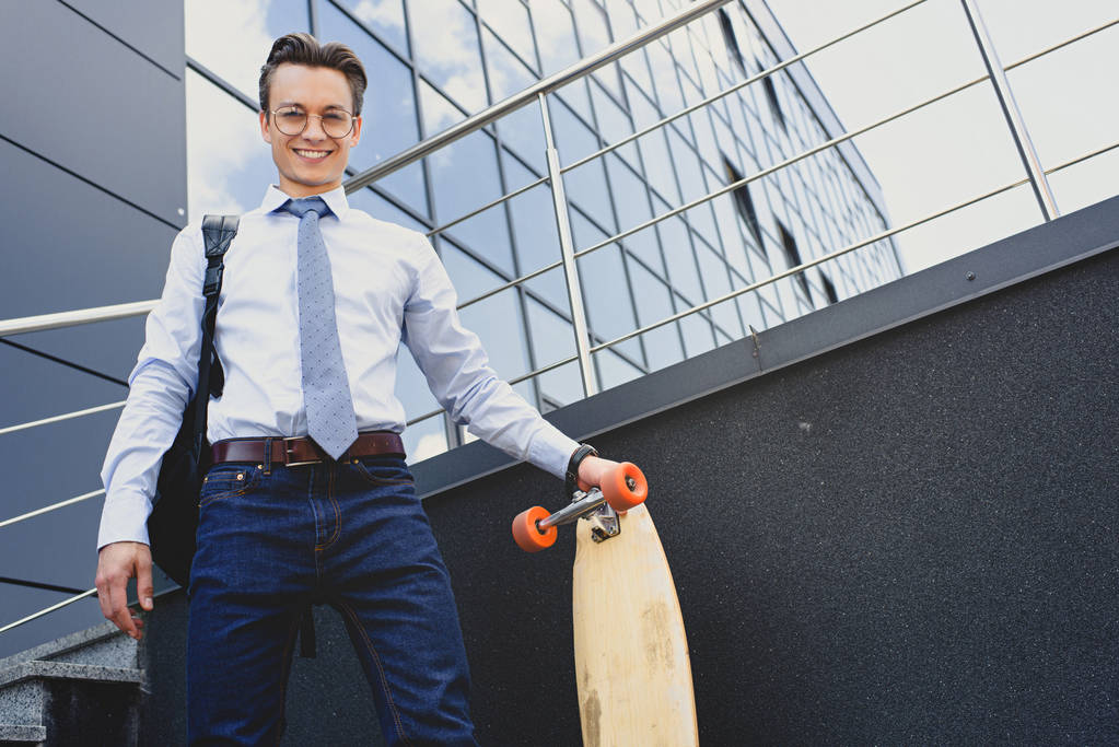χαμηλή γωνία προβολή του ωραίος νεαρός επιχειρηματίας σε γυαλιά στέκεται με longboard και να χαμογελά στη φωτογραφική μηχανή - Φωτογραφία, εικόνα
