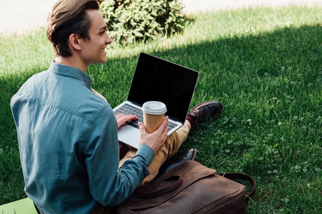 souriant jeune pigiste tenant tasse en papier et en utilisant un ordinateur portable avec écran blanc tout en étant assis sur l'herbe
 - Photo, image