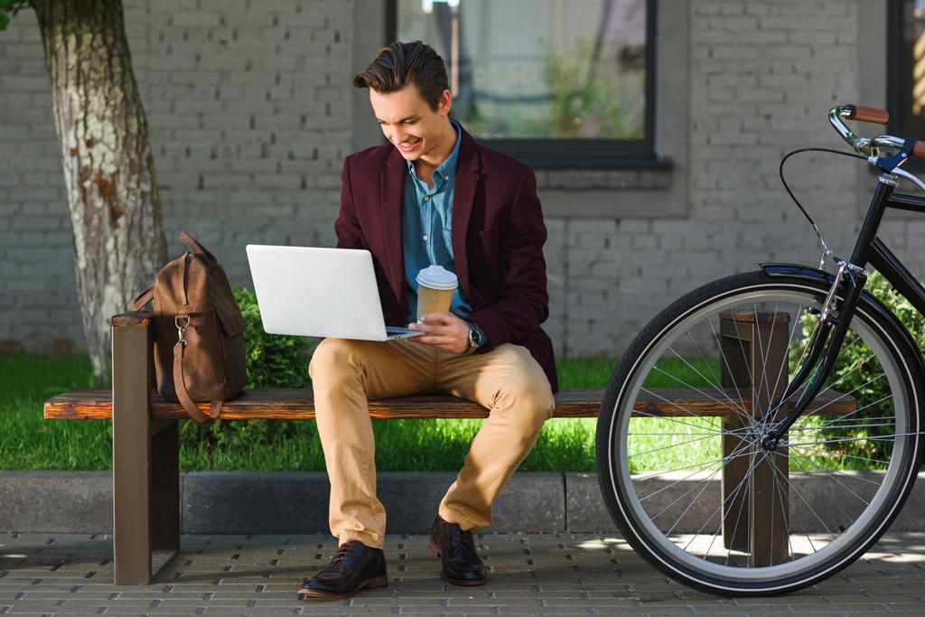 sorrindo jovem freelancer segurando copo de papel e usando laptop enquanto sentado no banco
 - Foto, Imagem
