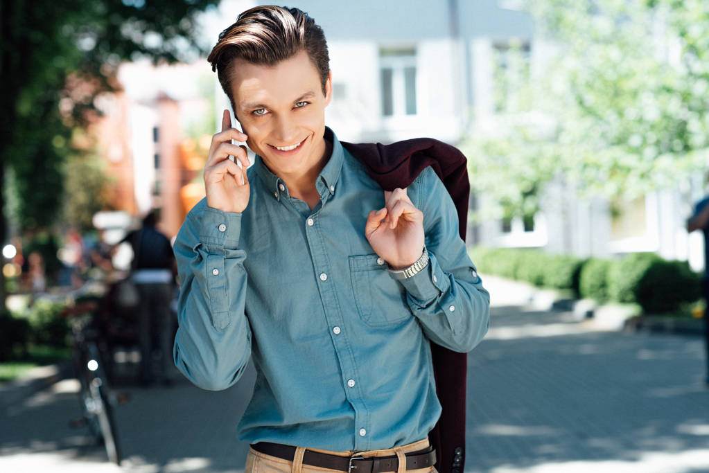 csinos, fiatal férfi beszél a smartphone és mosolyogva kamera utcában - Fotó, kép