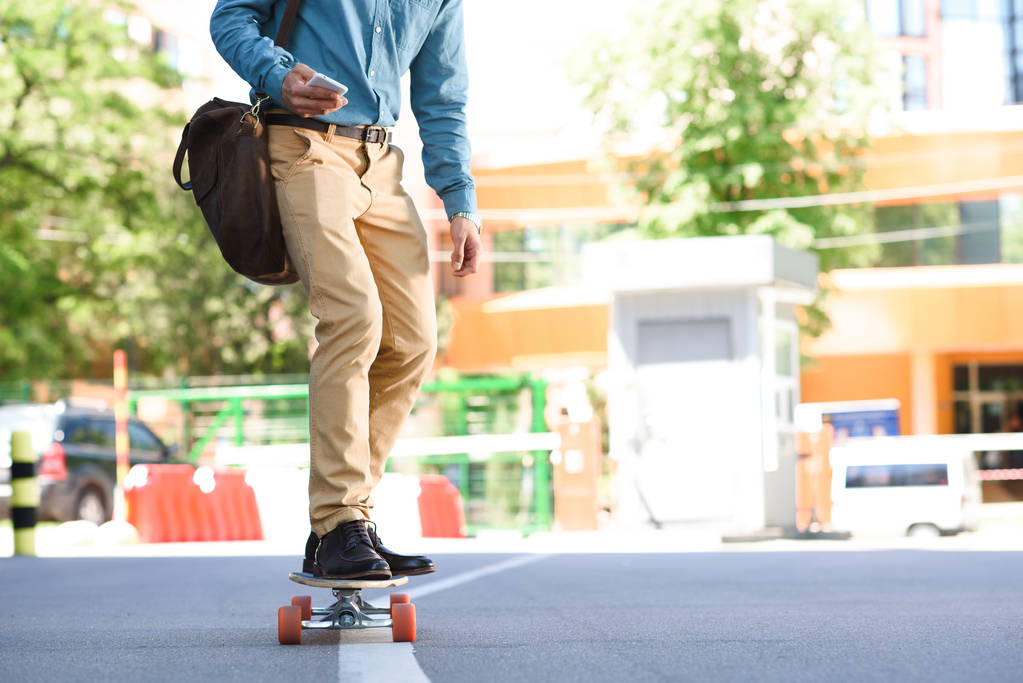 περικοπεί shot νεαρού χρησιμοποιώντας smartphone και ιππασία skateboard στην οδό - Φωτογραφία, εικόνα