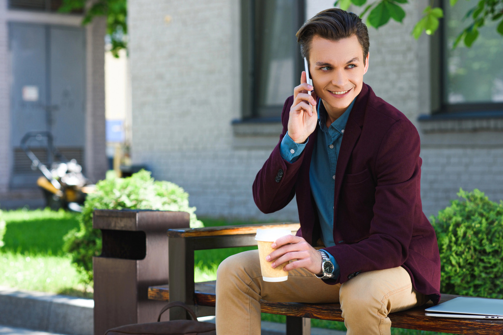 szép, mosolygós fiatalember gazdaság papír poharat, és beszél a smartphone-padon ülve  - Fotó, kép