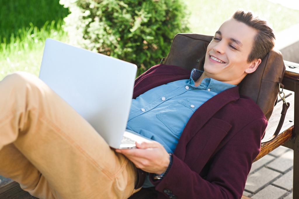 счастливый молодой фрилансер, лежащий на скамейке и использующий ноутбук
 - Фото, изображение