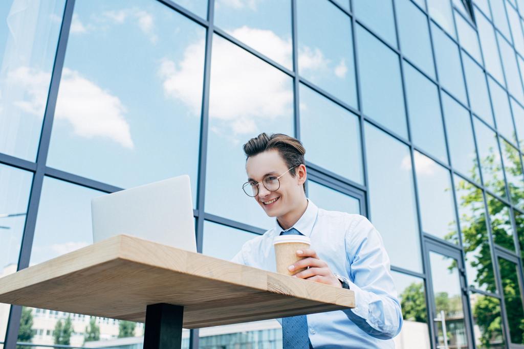 низький кут зору усміхненого молодого фрілансера, який тримає паперову чашку і використовує ноутбук за межами сучасної будівлі
 - Фото, зображення