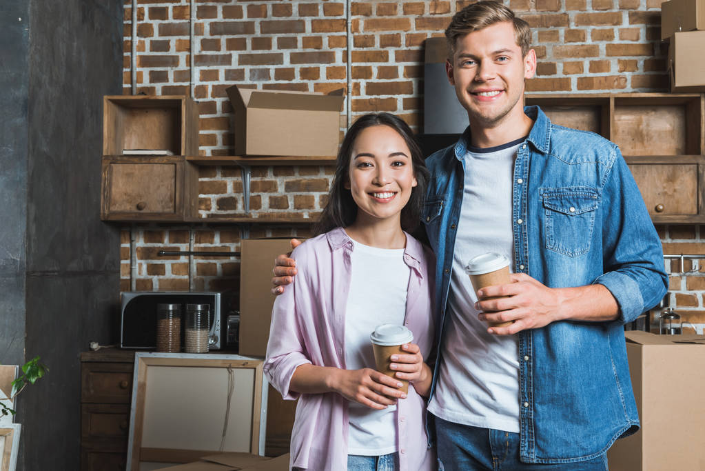 young interracial couple with paper cups of coffee standing on kitchen while moving into new home - Photo, Image