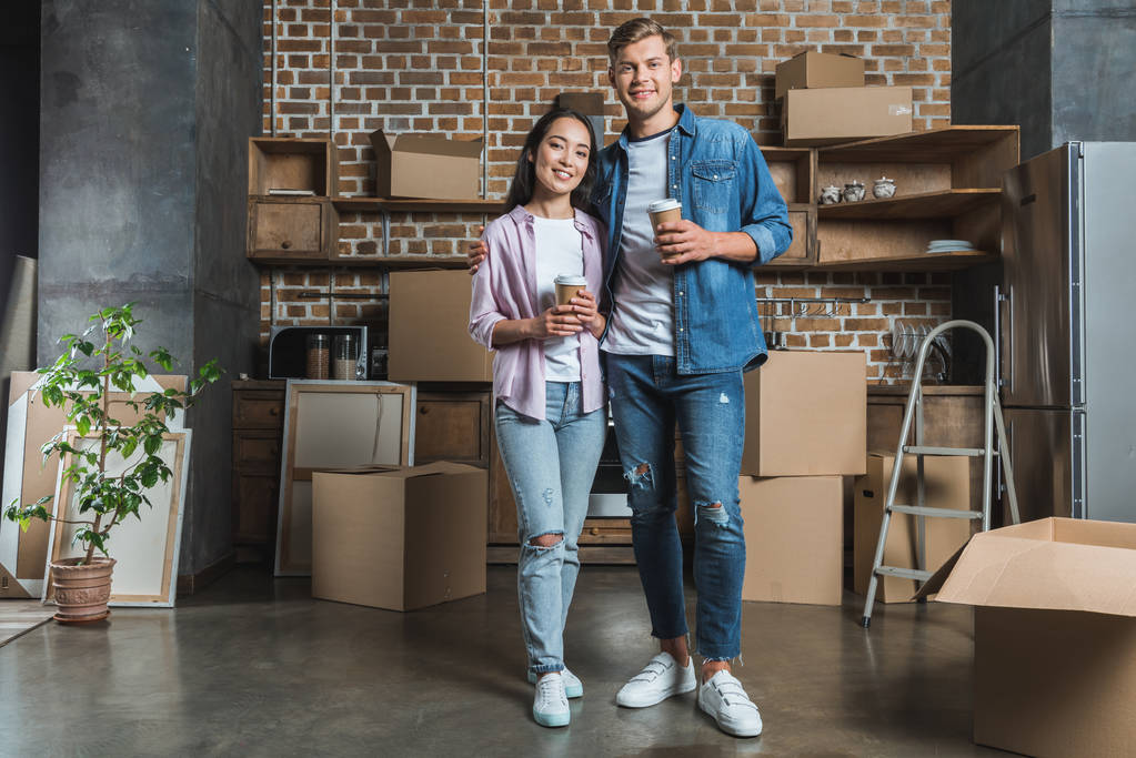 счастливая молодая пара с бумажными чашками кофе стоя на кухне, переезжая в новый дом
 - Фото, изображение