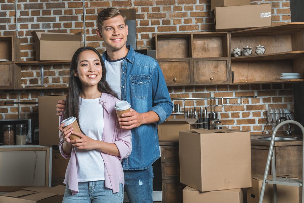 beautiful young couple with paper cups of coffee standing on kitchen while moving into new home - Photo, Image
