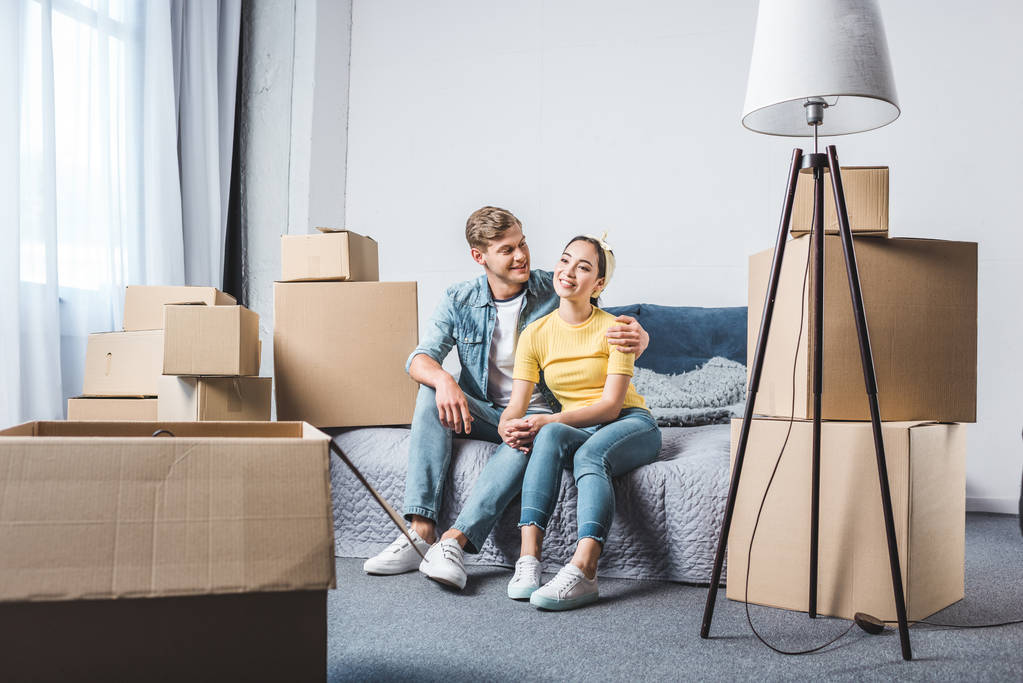 beautiful young couple sitting on bed after moving into new home surrounded with boxes - Photo, Image