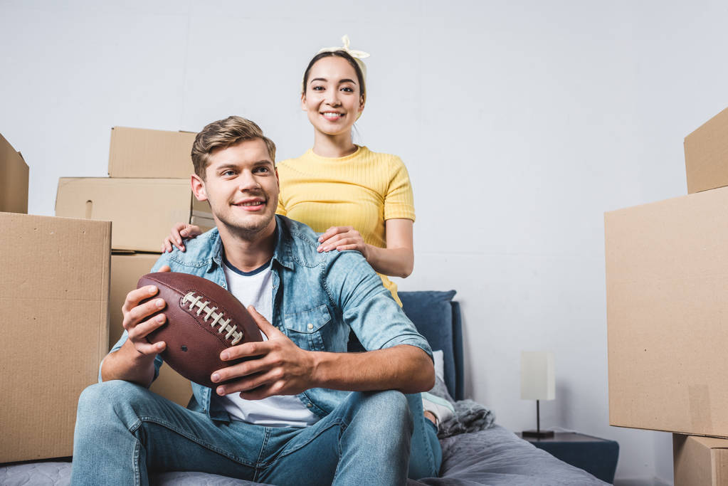 rotujenvälinen nuori pari istuu sängyllä amerikkalaisen jalkapallon kanssa muutettuaan uuteen kotiin
 - Valokuva, kuva